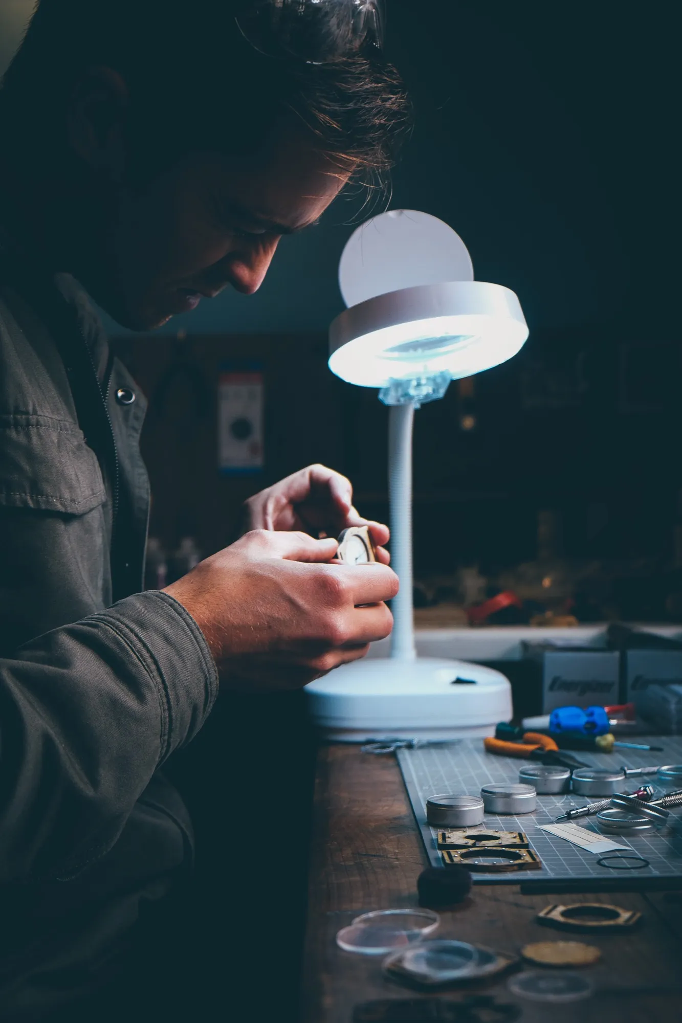 A craftsman examines a small watch closely under a desk lamp, with watch parts and tins on the table nearby