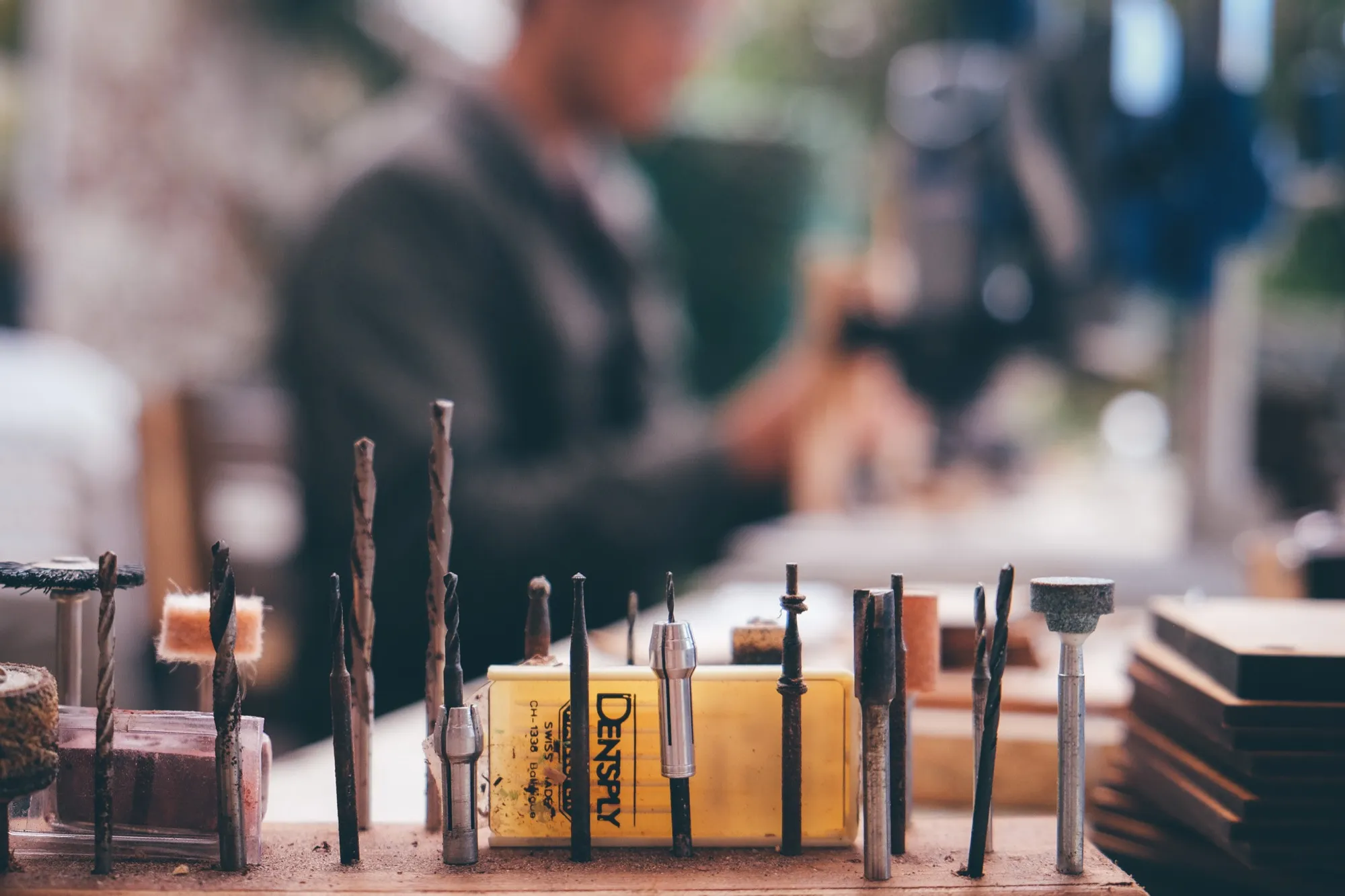 Rows of drill bits and small cutting tools standing upright on a workbench, with someone working blurred in the background