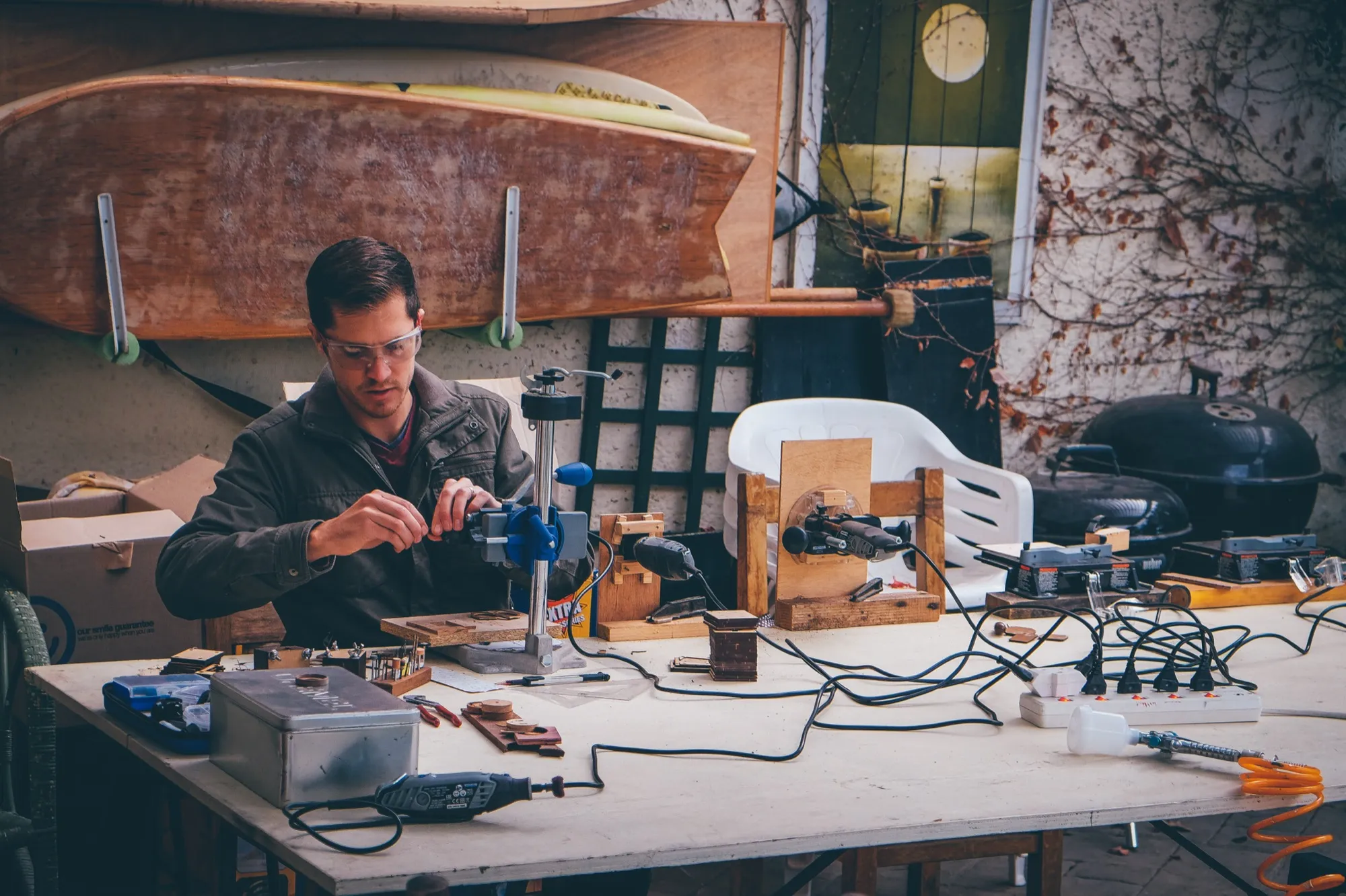 A craftsman in safety glasses shapes a small block of wood clamped in a rotary tool stand at an outdoor workbench
